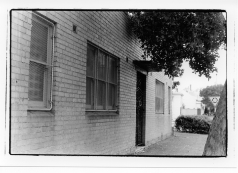 Black & white photo showing building entrance and brick wall as well as security wiring on windows. 