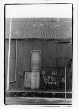 Black & white photo of a corrugated iron building with graffiti painted on side of wall.