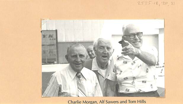 Three men in a casually posed photograph. The man on the right (Tom Hills) is smiling and pointing at someone behind the camera, perhaps the photographer.