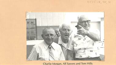 Three men in a casually posed photograph. The man on the right (Tom Hills) is smiling and pointing at someone behind the camera, perhaps the photographer.