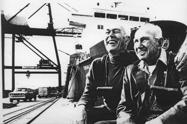 Black & white photo of two gentleman sitting on a pier in front of a large ship, a crane and vehicles  can be in the background.