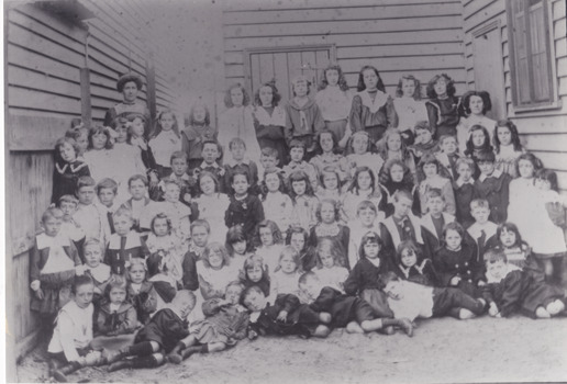 Black & White photo showing a large group of children dressed in Victorian clothing with an adult female standing at the back.