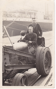 Man sitting, driving a tractor along a pier with another man standing on the back.