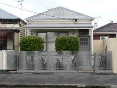 Small grey and white single fronted weatherboard cottage with a grey picket fence with 2 small gates, one leading up to the front door, and the other path to the side of the house, behind the fence there are two small shrubs and other plants. A large 6 panel window with grey painted edges, the front door is painted grey and a carved wood panel is 3/4 of the front door surface and above that area a panel contains eight small window panels.  
