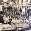 A large group of working celebrating Christmas, mainly women shown with a few males standing at the back of the group. Christmas decorations are hung along the walls and are on the table amongst their work area. 