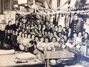 A large group of working celebrating Christmas, mainly women shown with a few males standing at the back of the group. Christmas decorations are hung along the walls and are on the table amongst their work area. 