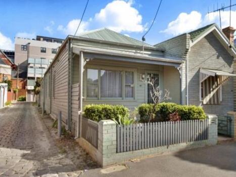 single fronted weathered board cottage with blue stone lane running along the left hand side of the house. The fence consists 3 small brick posts with picks ending running between post and  wrought iron gate.  The bricking and weather boards are painted light green.  There is a cement pathway leading unto the veranda and front door. There is a large window with 4 side panels with lace curtaining. A small garden with various plants is in the front of the house. In the background a new apartment building can be seen.