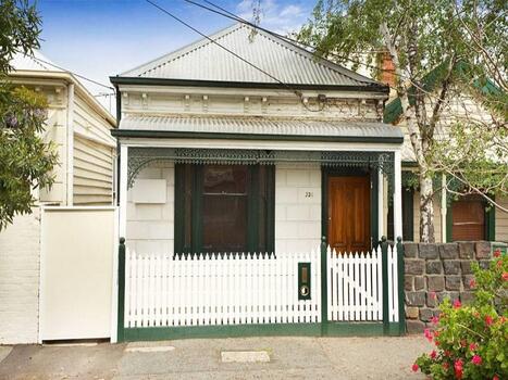 Small single fronted cottage painted creme, with bottle green paintwork around the window and door frame. There is a white picket fence with bottle green edging along the bottom of the fence, as well as the four fence posts and letter box.  The guttering and iron lace work are also painted. bottle green. The roofing is corrugated tin.   