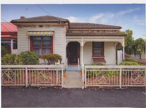  Double fronted creme coloured weathered board house. Cement pathway leads up to two steps onto veranda. Support rails are on either sides of the steps.There is a decorative white coloured wrought iron fence and gate with a garden on either side behind the fence with various plants. A wooden bench in on the veranda. On the left hand side of the house a 6 panel window with a red outer border and brown edge glass panels. A grey put down sun blind with yellow stripes its above the window. The veranda window has two panels of glass and the edge is also painted red. Iron lace work runs along the edge of the veranda roof between 3 poles and at the end of the veranda sonnets with the front wall of the house.  The roofing is brown curved ceramic tiles and a brown security door. 
