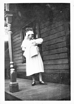 Woman in white holding a baby wrapped in a blanket on the verandah of a weatherboard house.