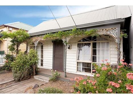 A white coloured double fronted white cottage with a pathway leading up two steps to a veranda. Two large windows with eight small panes of glass in each window are on the beachside of the house, the panes are painted creme and the four four ornate veranda posts are grey.  Iron lace roof goes between each of the grey posts and a vine of some sort is growing trough the iron lace work. There are various scrubs in the front garden.