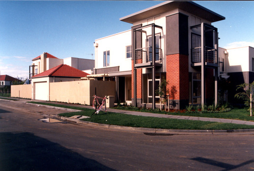 Newer style town house situated on a corner, large cream cement. A high fence with entrance to a garage runs along the side of the property. Small lawn and garden are in the front and side of house.