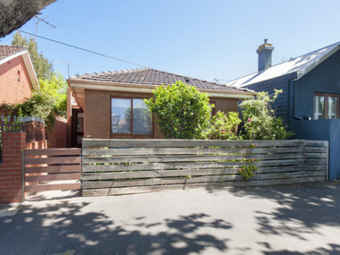 Newer style brick house with a tiled roof, side gate and main entrance on the left hand side of house. Two windows in the front and a small garden. High wooden unpainted front fence.