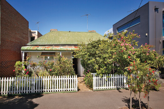 Double fronted weatherboard cottage situated between two brick buildings. Roof is made of corrugated iron, the outside of the cottage is painted white and there is a garden in front of the house contains various plants. There is a white Pickett fence and a brick pathway leading up to the veranda.  In the footpath in front of the cottage there is a small red bottlebrush tree.