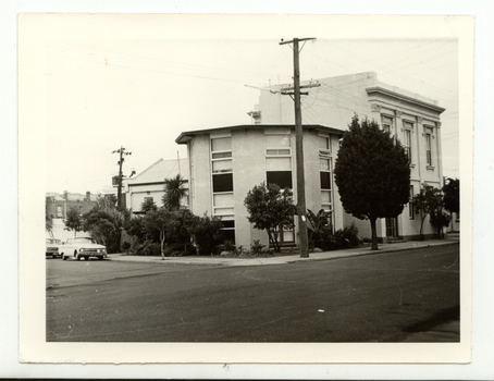 Light coloured rounded brick apartments situated on a corner, with large widows. Next to the rounded building there is larger double storey house also with large windows. Trees and shrubs  are om the footpath and the edge of the buildings. A car is parked to the side of the rounded building. 