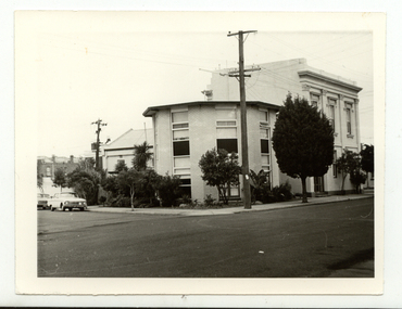 Light coloured rounded brick apartments situated on a corner, with large widows. Next to the rounded building there is larger double storey house also with large windows. Trees and shrubs  are om the footpath and the edge of the buildings. A car is parked to the side of the rounded building. 