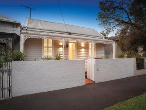 Double fronted creme coloured house, with medium size brick fence contains a small garden and a wooden gate.The weatherboard house has a corrugated tin roof and white iron lace work connects the four veranda posts. The front door, window frames ad gate are painted white. 