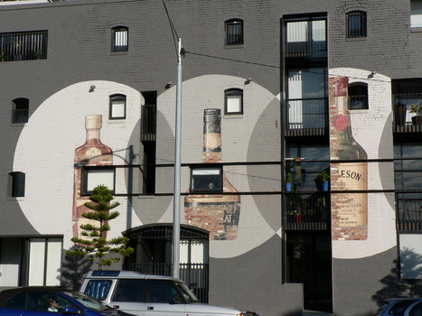 Images of 3 bottles of alcohol shown on the side of a redeveloped distillery. All images are contained within 3 white bubbles painted onto on a grey wall.  Several small windows and doorway are situation amongst the mural.