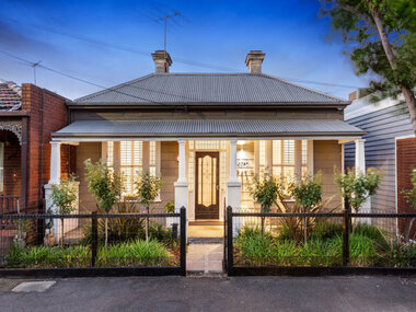 Double fronted coffee coloured house with small garden contains various plants in the front and a hooped wire fence and gate. Six pavers surrounded by small white pebbles lead up to the veranda.  The front door has a glass inset within a brown wooden frame with a ornate wire door, There is a glass panel above the front door and a glass panel on either side of the doorway. The window frames are painted white with four long panels of glass in each window. Four white wooden pillars line the veranda edge. Behind the windows and the front door are security bars.    