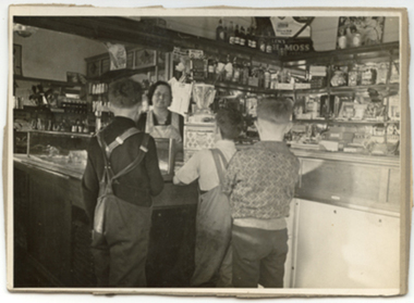 Three young boys dressed in dungarees and jumpers waiting to be served by an older lady wearing a pinafore and glasses in an old fashion milk bar. The shelves are stocked with various items for sale. 