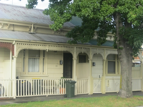 Two creme coloured single fronted attached cottages, both have separated ring panel fences with gates. Both have iron lace work on the edge of the roof below the guttering. Both have tin roofs. A large tree stands in the front of the cottages. 