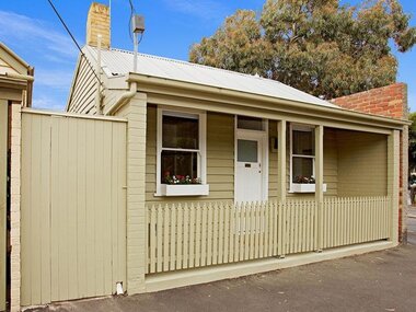 Latte coloured double fronted weather board cottage with a Pickett fence and side gate entrance. Front door and window frames are painted white with flower boxes attached to each window. Corrugated iron roof with a brick chimney. 