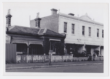An older style brick home with iron lace work and an unpainted wooden fence alongside three modern white coloured two story homes. All the homes have some shrubs in their front area. Port Melbourne, c. 1976