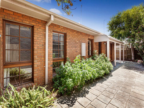 Light brown brick Unit with creme coloured guttering, downpipes and veranda roof. Top part of windows contain six small squares of glass and the lower part has four squares of glass the window frames are painted light brown. A selection of shrubs lined the front of the unit and a tree is to the side of the unit. The front area is paved with cement pavers.  c March 2015