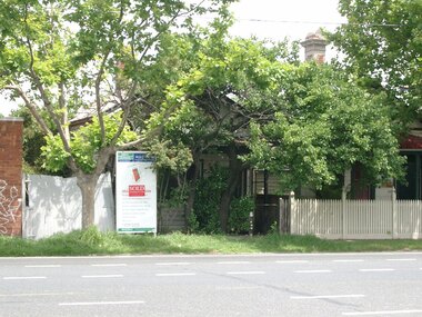 Large tree hides property up for auction, there is a auction board with a white fence to the left hand side of the auction board. The next door property has a cream coloured wooden fence. Another tree is situated on the grass nature strip in front of the house up for auction.