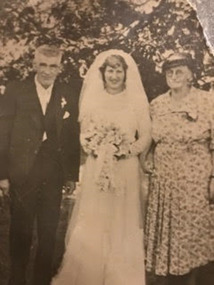 Two older persons standing alongside bride in a traditional white bridal dress with veil and bouquet of flowers. Gentleman is dressed in a suit and the lady is in a patterned dress and is wearing a darker coloured hat.