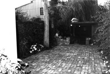 Small bricked yard with greenery along both fences and a few shrubs along the edge of the fences. Part of a large tree is shown and a partly built brick barbecue, there is a doorway leading into the building with tree branches on part of the doorway roof. c. 1980s
