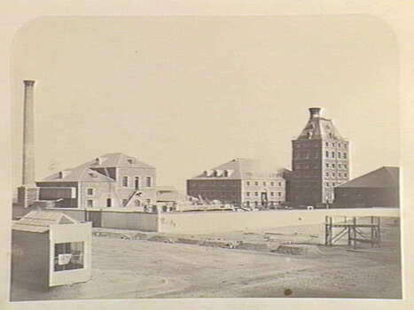 Photo shows the general layout of the buildings at the Sugar Works in Sandridge, c. 1860. There are 5 buildings shown in the photo as well as a large chimney at the side of one of the buildings. 