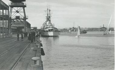 The view along Station Pier looking towards the shore with a few people standing on the pier and a warship berthed closer to shore. A few sailing yachts are also shown in the vicinity of the warship.