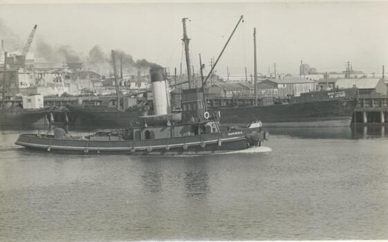 Photo shows the tug boat 'Marimba'  steaming past a wooden ship berth in the background, also shows a crane and wooden buildings in the background.