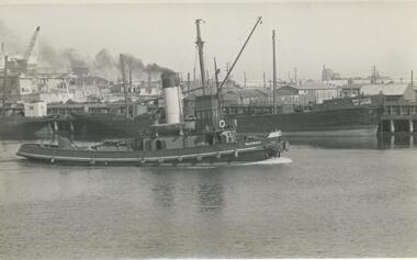 Photo shows the tug boat 'Marimba'  steaming past a wooden ship berth in the background, also shows a crane and wooden buildings in the background.