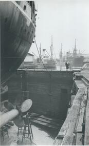 Photo shows part of a ship in the Duke's and Orr's dry dock facing the gate at river end. In the background you can see numerous ships berthed alone the river.