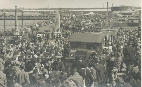 Photo shows people standing on Centenary Bridge,  Port Melbourne. Ambulances are carrying wounded soldiers back from World War 11. In the background you can see more people lining the old pier and the road leading up to the Centenary badge
