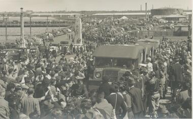 Photo shows people standing on Centenary Bridge,  Port Melbourne. Ambulances are carrying wounded soldiers back from World War II. In the background you can see more people lining the old pier and the road leading up to the Centenary badge