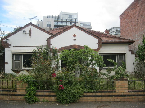 The front facade, garden and wall of white house with red brick decorative elements and a tiled roof. The top of a tall apartment building is visible in the background