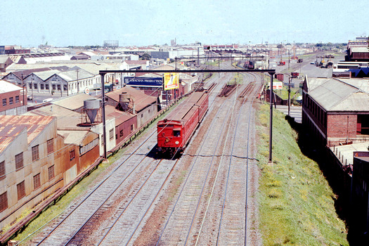 A red coloured train on railway tracks laid through an industrial area viewed from a raised position.