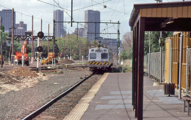 A silver coloured train approaches an empty platform with the tall buildings of the city in the background.