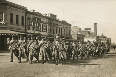 A group of soldiers in uniform carrying their overcoats marching along a street with two storey buildings in the background.