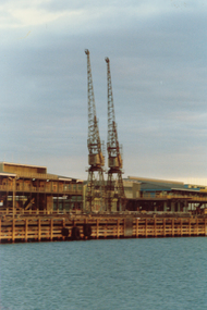Two large cranes on a wharf situated  in-between wharf buildings. 
