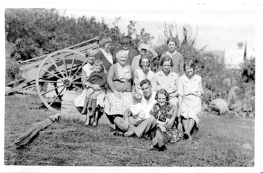 Group of thirteen men, women and children posing near an empty two-wheeled cart. 