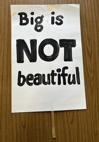 White cardboard placard attached to a wooden handle with black hand-printed slogan - Big is NOT beautiful