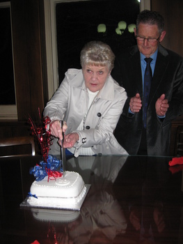 A woman wearing a silver jacket cuts a cake decorated with red and blue ribbons while a man watches on applauding.