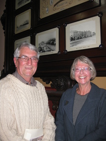 A  man and a women smile for the camera. A collection of historical photos are on the wall behind them.