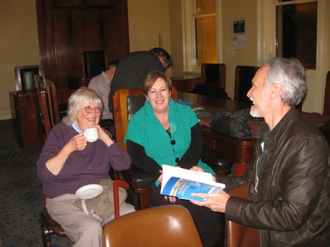The woman on the left is drinking from a cup and saucer while the woman next to her smiles towards the camera. A man side on to the camera is holding a book and smiling towards the two women.