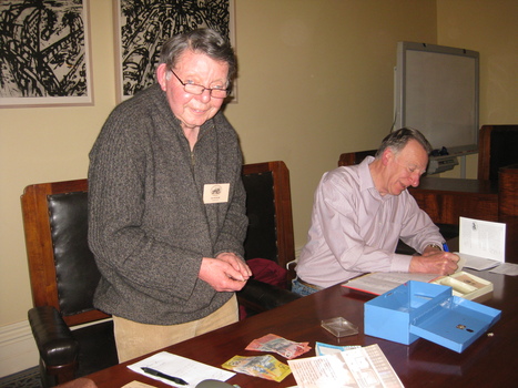 A man stands with papers and money, $50 and $20 notes, and an open cash tin on the table in front of him. A second man, sitting at the table, is writing something in a book.