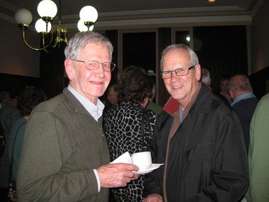 Two men are standing in a crowded room, the one on the left is holding a cup and saucer, both smile towards the camera.